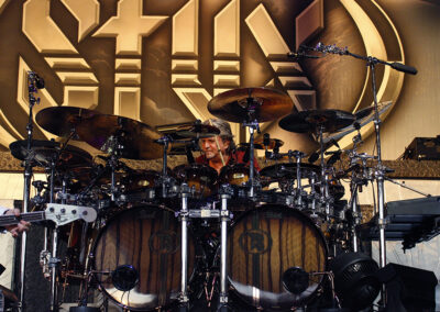 A drummer surrounded by an elaborate drum kit performs on stage, with large brown drums and numerous cymbals. The backdrop features a large stylized “STYX” logo in gold and silver tones.