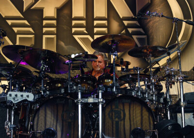 A drummer sits behind an elaborate drum kit on stage, surrounded by multiple cymbals and drums, with a large band logo in the background.