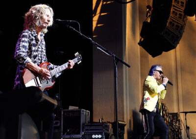 Two musicians perform on stage: one plays electric guitar and sings into a microphone, while the other, wearing sunglasses and a yellow jacket, sings passionately. Stage equipment and lighting are visible in the background.