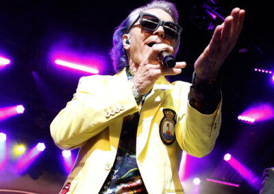 A person with gray hair wearing sunglasses, a bright yellow jacket, and a colorful shirt sings into a microphone on stage under vibrant pink and purple lights.