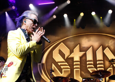 A singer with blue-gray hair, sunglasses, and a yellow jacket performs passionately on stage, holding a microphone. A large STYX logo is illuminated in the background amid colorful stage lights.