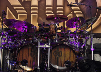 A drummer plays a large drum kit with multiple cymbals and drums, lit by purple stage lights. A stylized band logo is visible in the background.