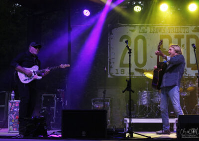 Two musicians perform on stage under colorful lights; one plays electric guitar, the other acoustic, in front of a Zac Brown Tribute sign. Drums and equipment are visible in the background.