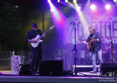 Two musicians perform on an outdoor stage under colorful lights. One plays an electric guitar, the other strums an acoustic guitar and sings. A drum set and equipment are visible in the background with a “20 RIDE” banner hanging behind them.