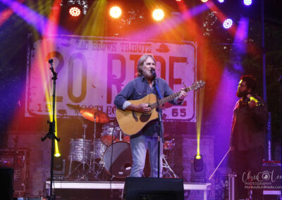 A musician plays an acoustic guitar and sings on stage under colorful lights, with a Zac Brown Tribute banner in the background and a band member playing bass to the right.