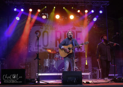 A musician plays an acoustic guitar and sings on an outdoor stage with colorful lights. A second performer stands nearby with a microphone. The backdrop reads “Zac Brown Tribute: 20 RIDE.” Drums and other instruments are visible.