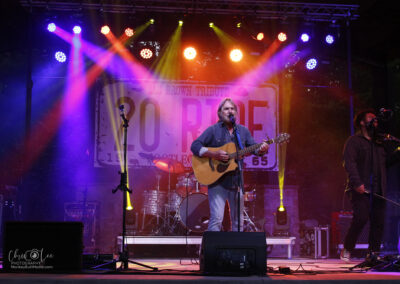 A male musician plays an acoustic guitar and sings on an outdoor stage with colorful lights, while another band member stands beside him with a microphone. A large “SO RIDE” banner hangs in the background.