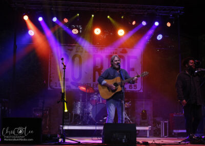 A musician plays an acoustic guitar and sings on an outdoor stage with colorful lights. A second performer stands to the right with a microphone. A drum set and amplifiers are in the background under a large ROAD sign.