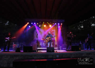 A band performs on an outdoor stage at dusk under colorful lights. The lead singer plays guitar center stage, flanked by musicians on electric guitar, drums, bass, and violin. A festival banner hangs in the background.