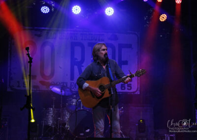 A musician with long hair and a beard plays an acoustic guitar and sings on stage under colorful lights, with a large 20 RIDE sign in the background and musical instruments around.