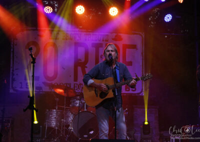 A musician with long hair plays an acoustic guitar and sings into a microphone on a colorful, spotlight-lit stage, with a drum set and a large “30 RIDE” sign in the background.