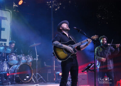 A bearded musician in a hat passionately sings and plays guitar on stage, accompanied by a drummer and a double bassist, with colorful stage lights and smoke effects highlighting the performance.