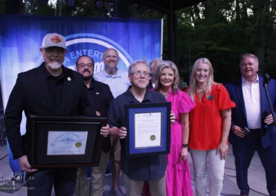 A group of people pose together outdoors; one person holds a framed certificate, and another holds a document. A blue event backdrop and trees are visible in the background.