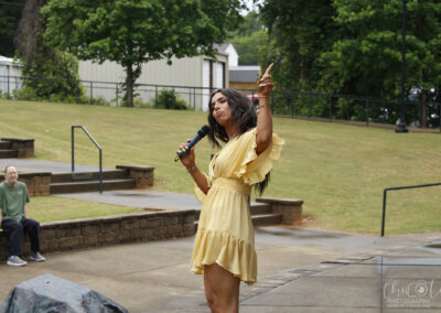 Woman in a yellow dress sings into a handheld microphone outdoors, lifting one arm in a performance pose as a man sits on stone steps nearby.