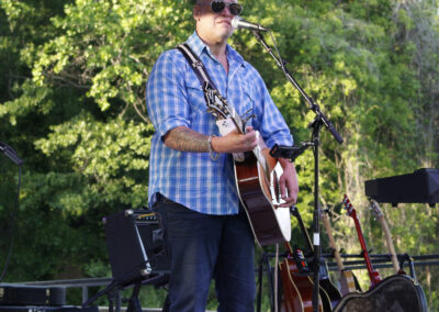 Male guitarist singing into a microphone on an outdoor stage, wearing a blue plaid shirt and sunglasses with green trees in the background.