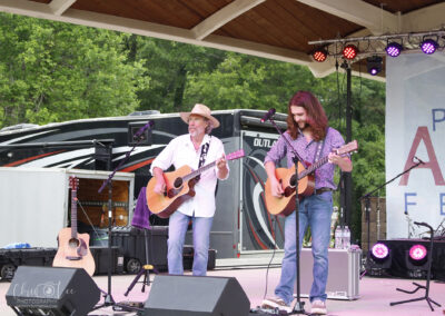 Two musicians play acoustic guitars and sing on an outdoor stage under a canopy, with equipment, a tour bus, and trees in the background. Stage lights are on and there is a festival banner partially visible.