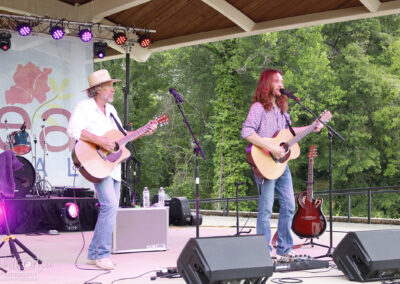 Two musicians with long hair play acoustic guitars and sing on an outdoor stage under a wooden roof. There are microphones, amplifiers, and a drum set in the background, with trees visible behind the stage.
