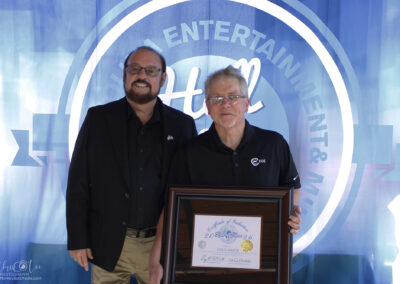 Two men stand smiling in front of a blue backdrop with a logo. The man on the right holds a framed certificate. Both are wearing dark-colored shirts and glasses.