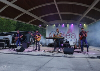 Four musicians perform on an outdoor stage under a wooden roof, playing guitars and singing. Purple lights shine, and smoke effects are present. A banner behind them reads Pickering and Art. Trees and equipment are visible.