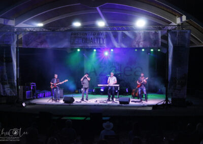 A four-member band performs on an outdoor stage at night under colorful lights, with musical instruments and equipment visible. A banner above them reads Presented by: EG Quality.