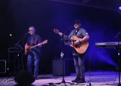 Two musicians perform on stage: one plays bass guitar and sings at a microphone, the other plays acoustic guitar and sings. Purple stage lighting and keyboards are visible in the background. Both wear glasses and casual clothing.