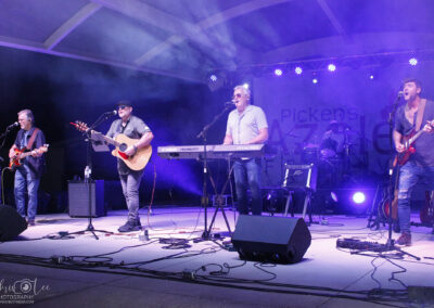 Five musicians perform on a stage with guitars, keyboard, and drums under purple lighting. They sing into microphones, and musical equipment surrounds them. A partially visible “Pickens… Jazz… Lee” banner hangs in the background.