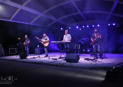 A five-member band performs on an outdoor stage at night, illuminated by purple and white lights. The musicians play guitar, keyboard, drums, and bass with a backdrop reading Pickens Azalea.