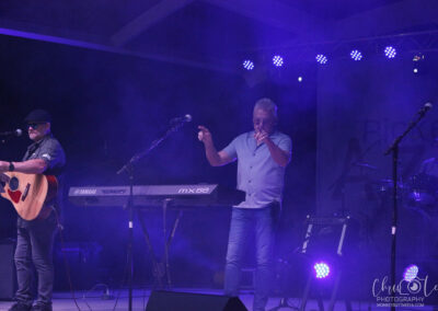 Two musicians perform on stage under purple-blue lights and fog; one plays guitar and sings, while the other stands at a keyboard and sings into a microphone. The stage has a Yamaha keyboard and a visible lighting rig above.