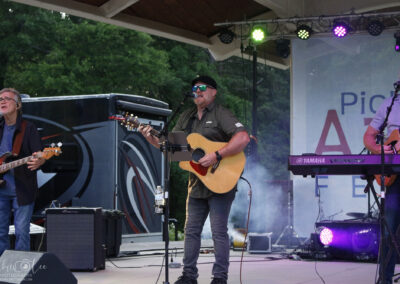 A band performs on an outdoor stage with guitars and a keyboard. Three musicians are visible, including a singer with a hat and sunglasses at the center. Trees and stage lights are in the background.