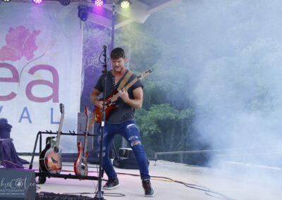 Male guitarist performing on an outdoor stage with fog and colored lights, microphone stand nearby.