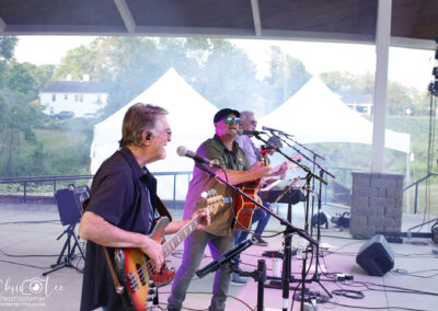 Three musicians perform on stage under a pavilion, playing guitars and singing into microphones. White tents and green trees are visible in the background during daylight. Sound equipment is set up around them.