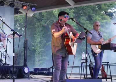 A band performs on an outdoor stage surrounded by trees. The lead singer plays acoustic guitar and sings into a microphone, while another musician plays keyboard. Stage lights and mist add to the lively atmosphere.