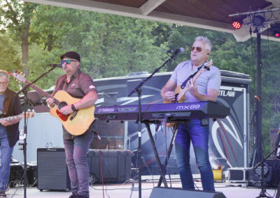 A band performs outdoors on a stage; one man plays acoustic guitar and sings into a microphone while another plays a keyboard and sings. Trees and an RV are in the background. The musicians wear sunglasses and casual clothes.