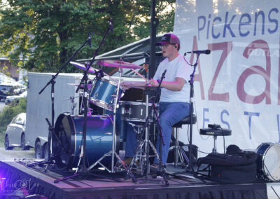 A man wearing sunglasses and a cap plays a blue drum set on an outdoor stage at the Pickens Azalea Festival, with microphones set up and trees and cars in the background.