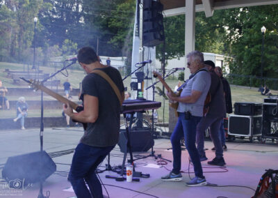 A band performs on an outdoor stage with electric guitars and microphones as people sit and watch on benches in the background; trees and equipment are visible around the stage.