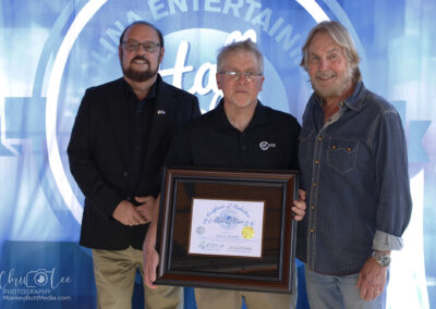 Three men stand in front of a blue backdrop. The man in the center holds a framed certificate. All three are smiling and dressed in casual or semi-formal attire. A photography watermark is visible in the corner.
