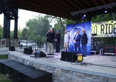 Five men stand on an outdoor stage under a pavilion, with microphones and stage lights. One man speaks into a microphone while others listen. A large 20 Ride tour banner hangs in the background. Trees are visible behind the stage.