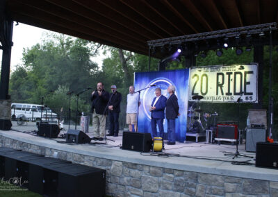 Five men stand on an outdoor stage under a wooden canopy, with microphones and musical equipment around them. A large 20 Ride tour sign and banners are displayed behind them. Trees are visible in the background.