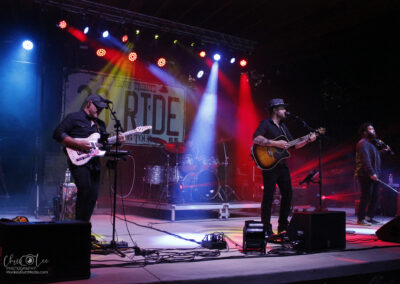 A live band performs on an outdoor stage at night, illuminated by colorful stage lights. Three musicians are at the front with guitars and microphones; drums and other instruments are visible in the background.