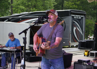 A man in a gray shirt and cap sings and plays an acoustic guitar on an outdoor stage, with another man playing a steel guitar nearby. An RV and trees are visible in the background.