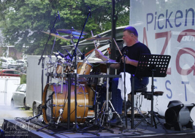 Drummer on a stage outdoors playing a large drum kit with microphones, wearing a black shirt and ear monitor, against a banner backdrop.