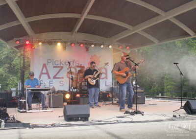 A country band performs on an outdoor stage at the Pickens Azalea Festival, with musicians playing guitar, drums, and keyboard under a wooden pavilion, surrounded by trees and stage lights.