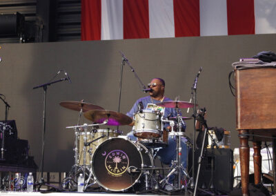 A drummer sings into a microphone while playing drums on stage during a live performance. There is a large drum with Down North written on it, and an American flag hangs in the background.