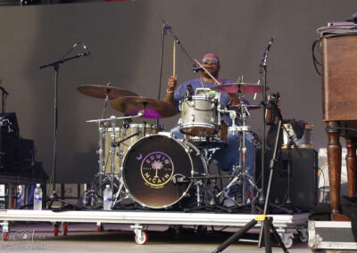 A drummer wearing sunglasses plays a drum kit on stage during a live performance, surrounded by microphones and music equipment, with the drum featuring a Edwin McCain logo on the front.