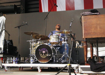 A drummer wearing sunglasses plays a drum set on stage, with microphones set up nearby. A U.S. flag is partially visible above, and a keyboard instrument is positioned to the right.