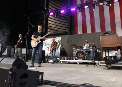 A band performs on an outdoor stage with American flag backdrop. Four musicians play guitar, bass, and drums, while purple lights shine overhead. Musical equipment and speakers are visible in the foreground.