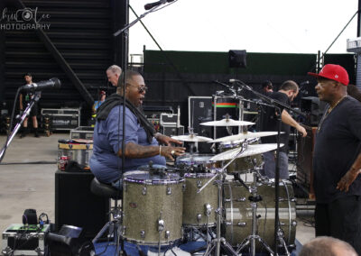 A drummer sits at a sparkling drum kit on an outdoor stage, energetically playing while talking to a man in a red cap. Crew members and equipment are visible in the background.