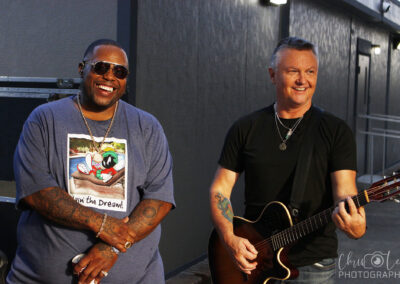 Two men are smiling outdoors; one wears sunglasses and a cartoon T-shirt, and the other holds an acoustic guitar, wearing a black shirt and necklace. A gray wall and some lights are in the background.