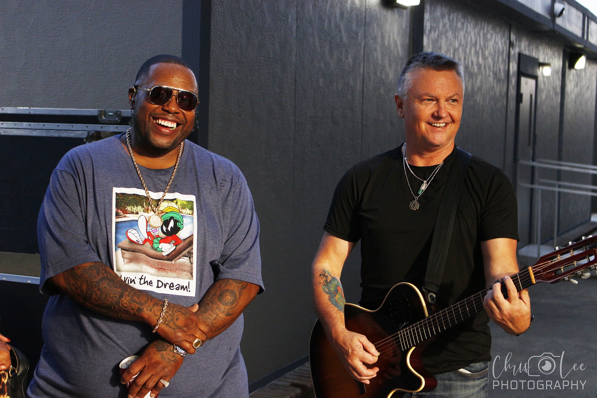 Two men are smiling outdoors; one wears sunglasses and a cartoon T-shirt, and the other holds an acoustic guitar, wearing a black shirt and necklace. A gray wall and some lights are in the background.