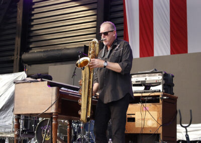 A musician wearing sunglasses plays a baritone saxophone on stage beside a wooden keyboard. Audio equipment and a drum set are visible in the background, along with a large American flag hanging above.